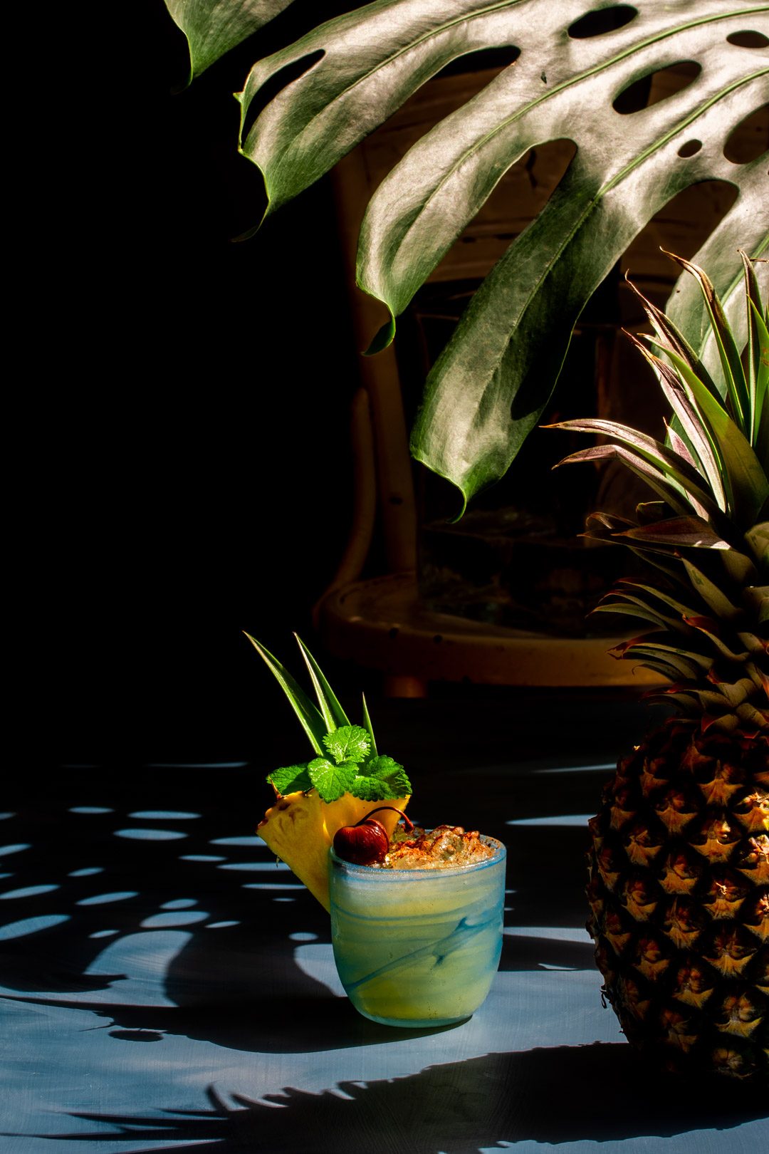 Pickle Mai Tai cocktail framed by shadows from a pineapple and monstera leaf