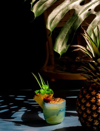 Pickle Mai Tai cocktail framed by shadows from a pineapple and monstera leaf
