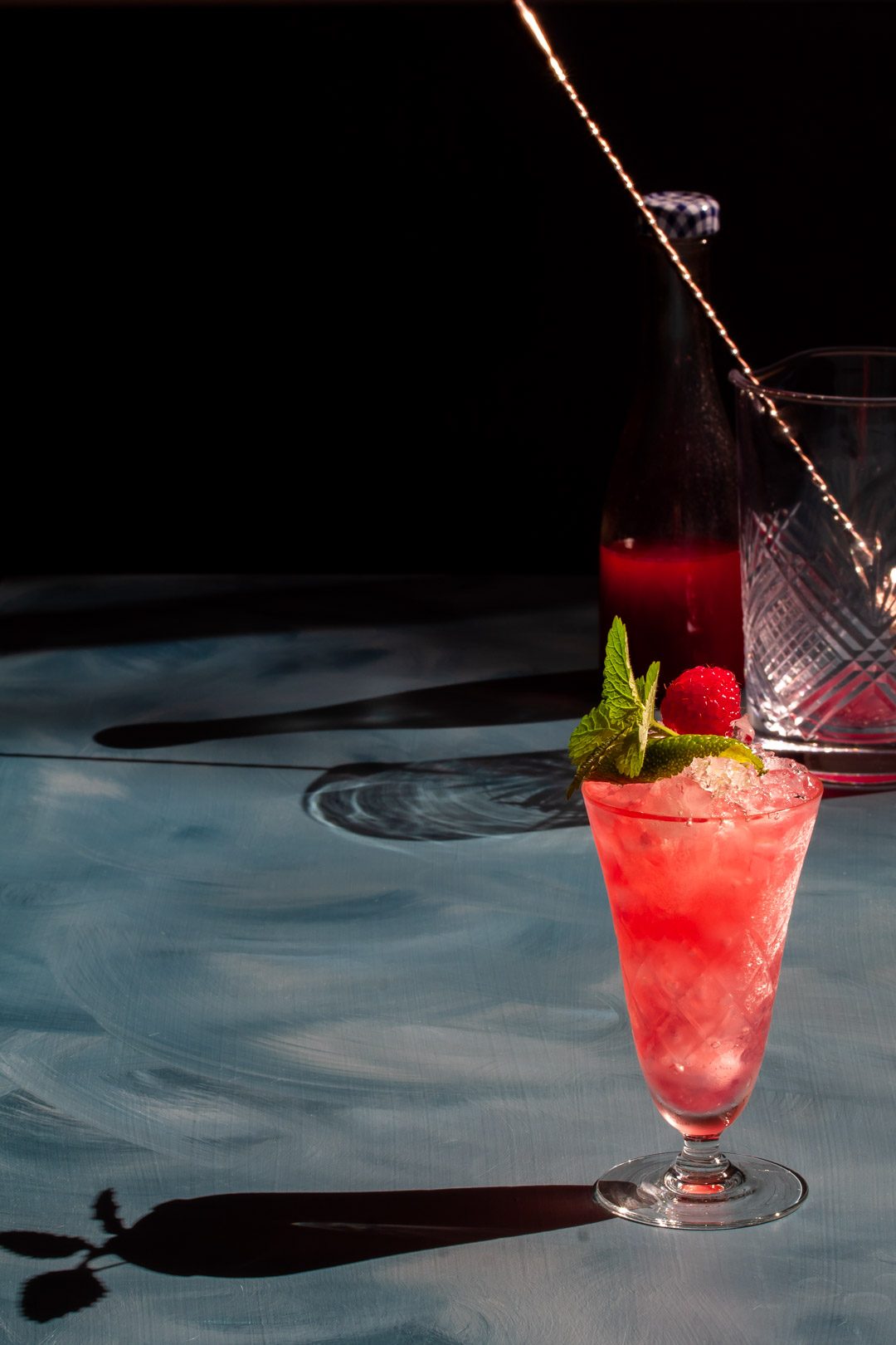 Making a Raspberry lime gin fix cocktail with shadow details, shrub bottle and mixing glass in background