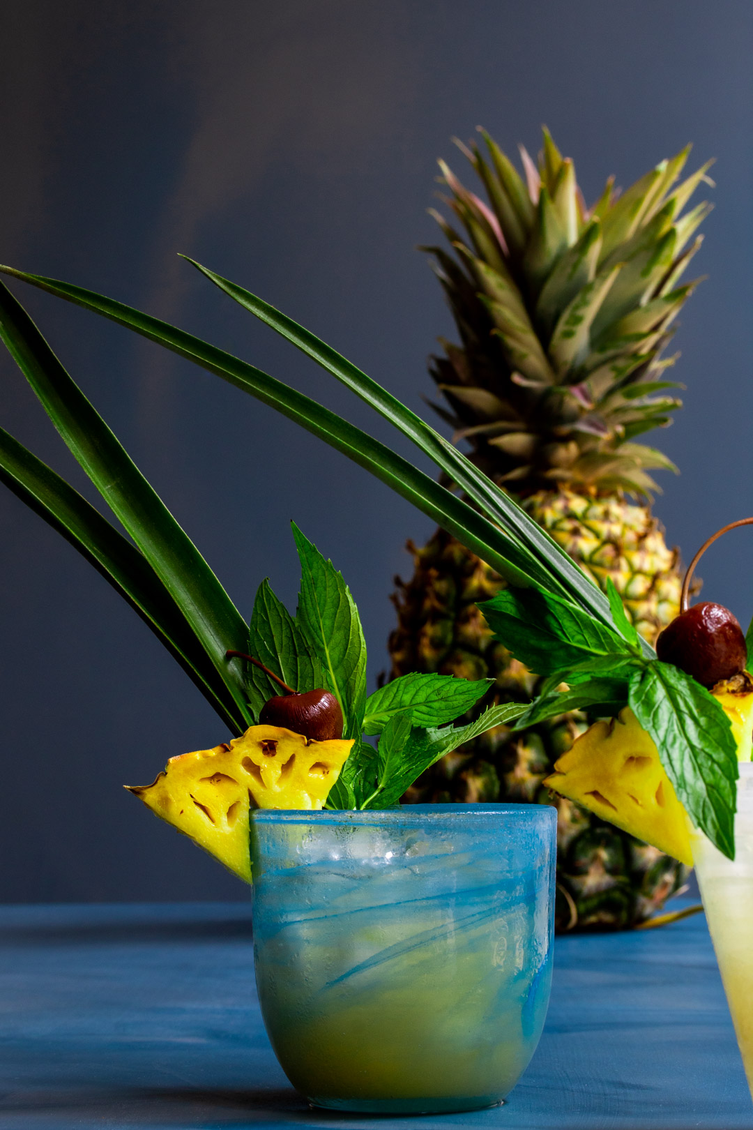 Pickle Mai Tai cocktail with blue glass front and pineapple in background