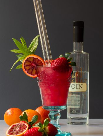 blood orange pomegranate gin daisy cocktail with bottle in background and fruit in foreground