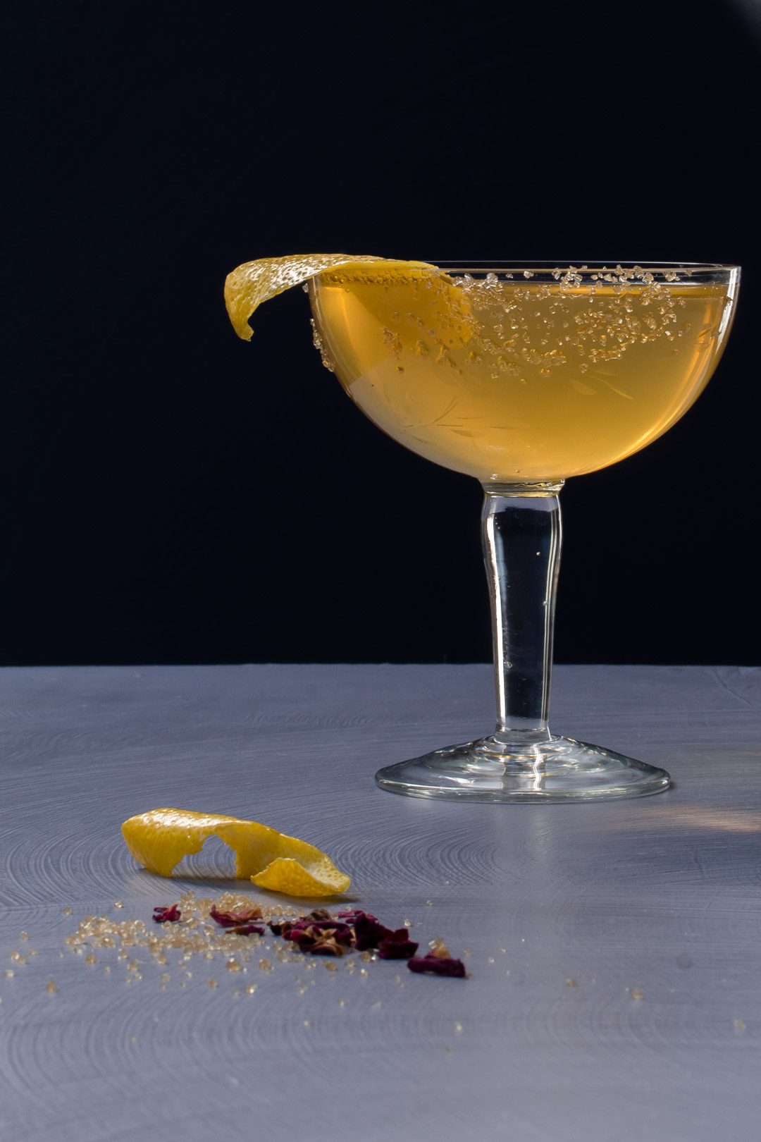 Fancy quince brandy champagne cocktail to left with lemon, rose petals, demerara sugar in right foreground