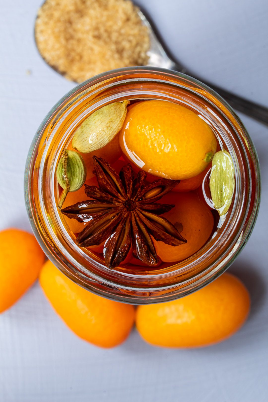 cumquat brandy from above with demerara sugar and 3 cumquats on light purple background