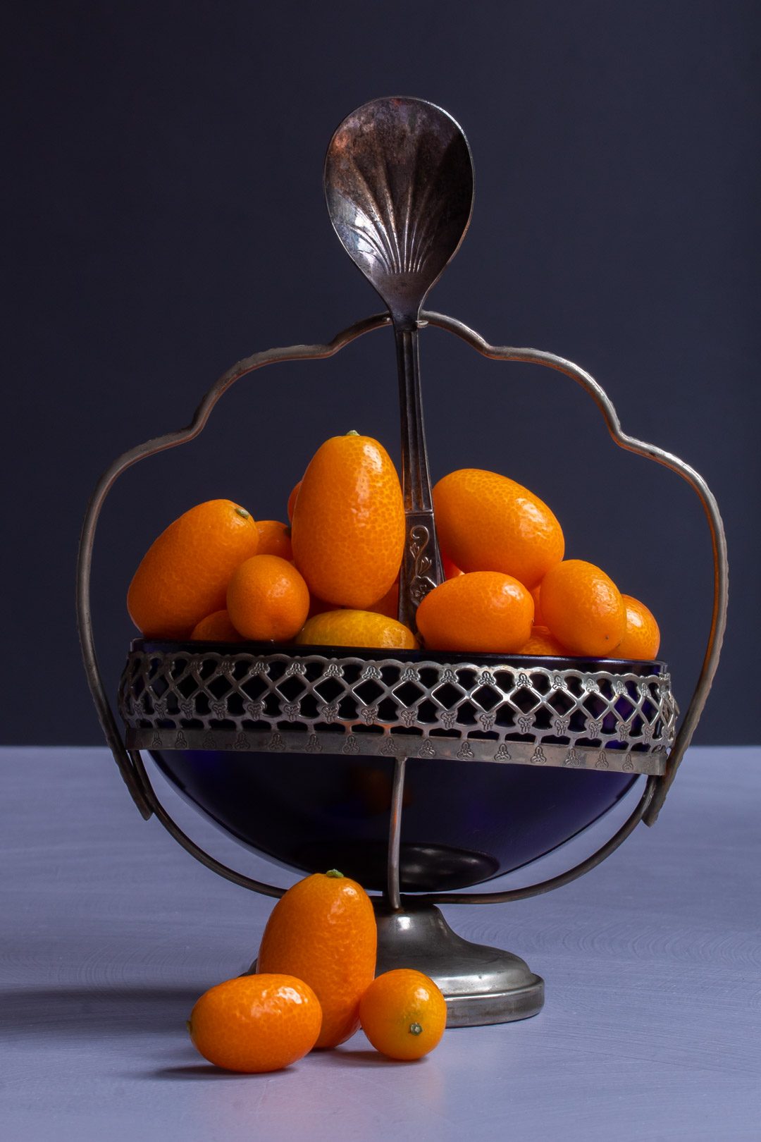 making cumquat brandy with cumquats in blue glass sugar bowl with 3 cumquats in foreground and sugar spoon on light purple and grey background