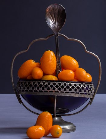 making cumquat brandy with cumquats in blue glass sugar bowl with 3 cumquats in foreground and sugar spoon on light purple and grey background