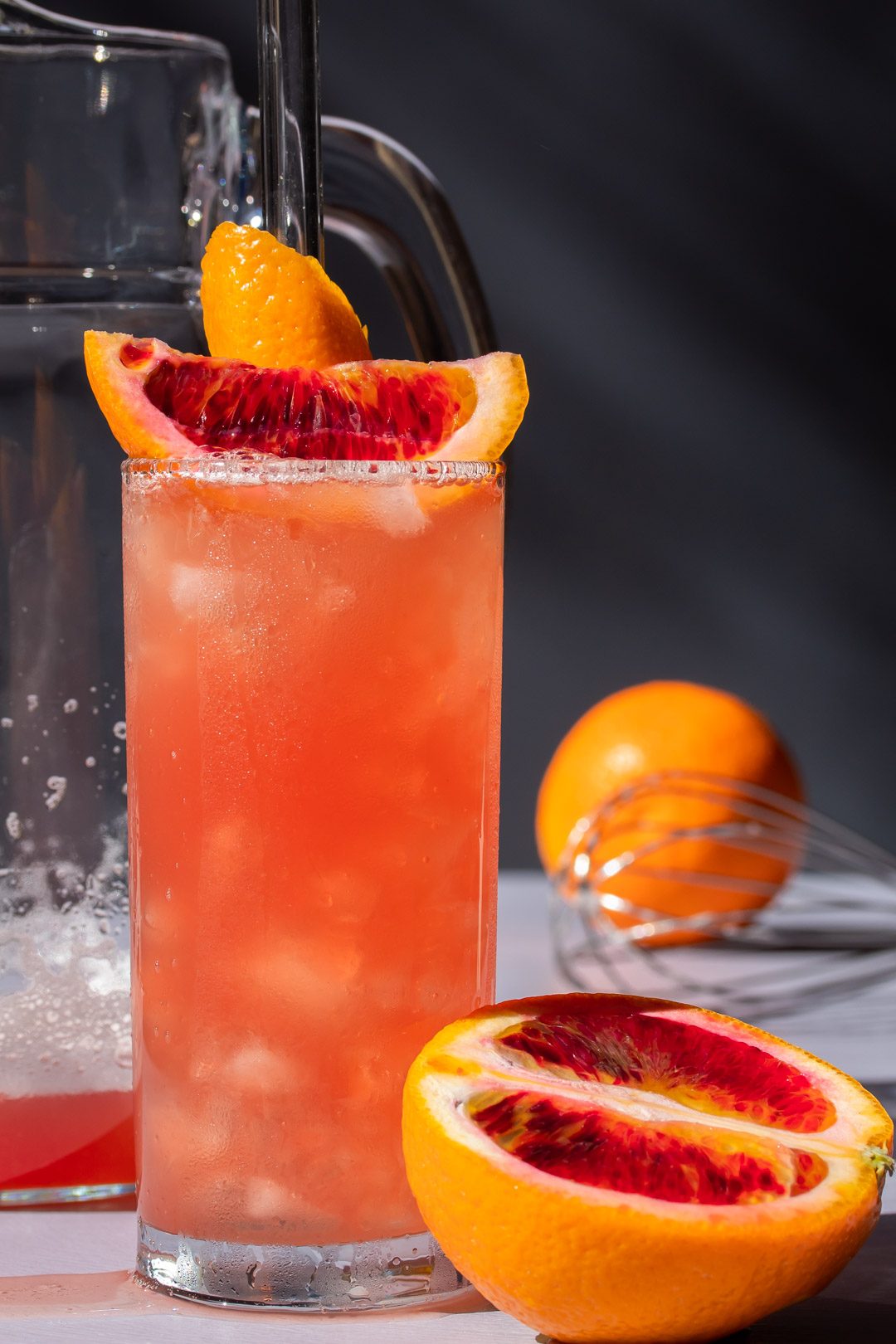 improved blood orangeade with jug, whisk and orange in background, cut blood orange in foreground