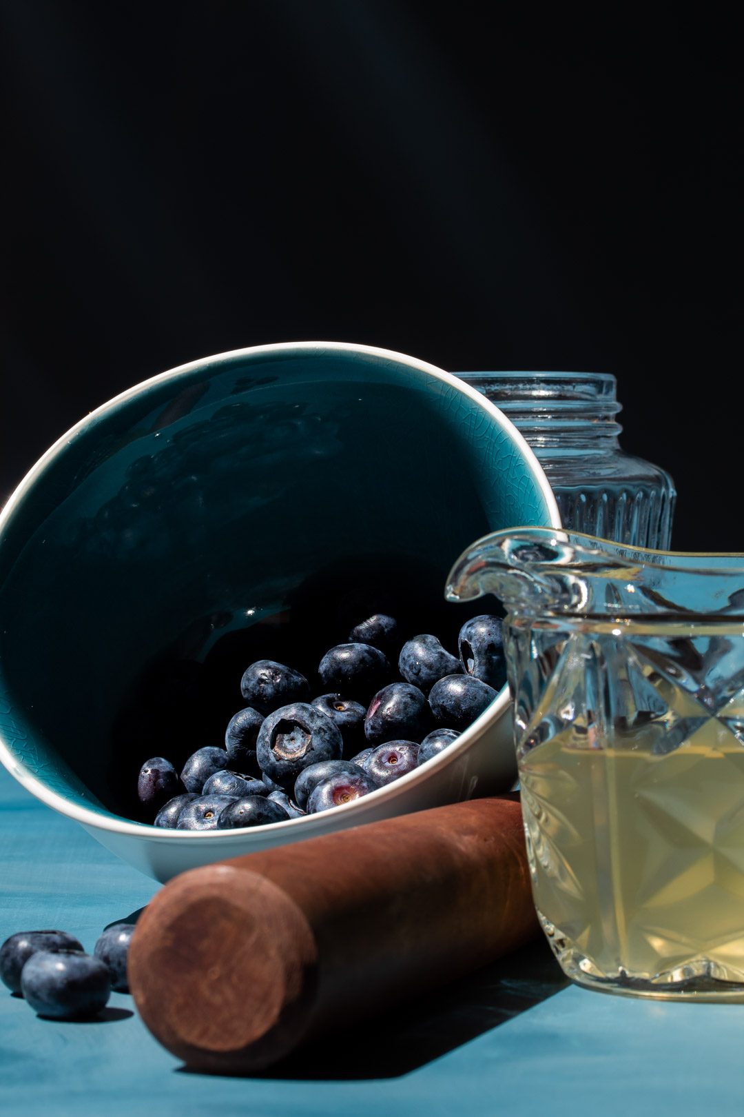 making blueberry shrub syrup with blue bowl of blueberries and cut glass vintage jug of coconut vinegar